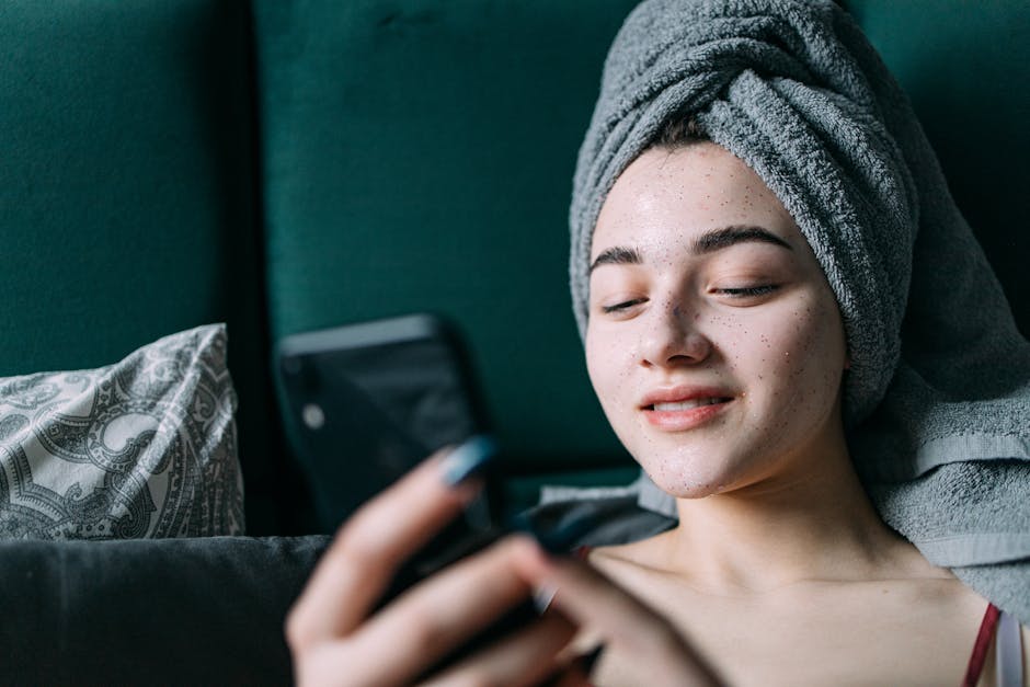 Young woman enjoying skincare routine, relaxed at home with a towel on her head, holding a smartphone.
