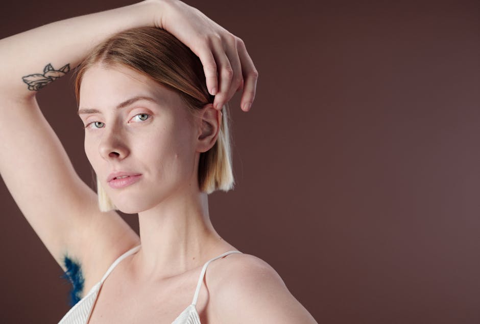 Close-up portrait of a young woman with an arm tattoo and blue underarm hair against a brown background.