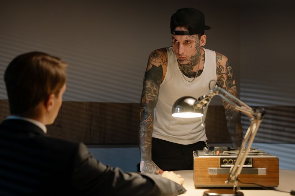 Tattooed man in a police interrogation room leaning over a desk with a tape recorder and lamp.