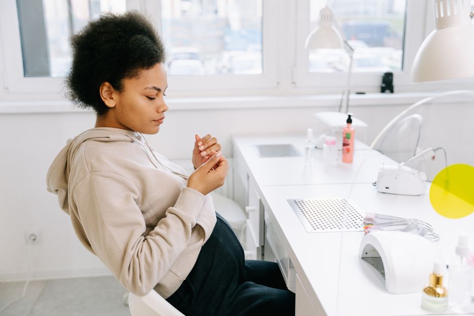 Pregnant woman with afro hair sitting at a nail salon, looking at her manicured nails.