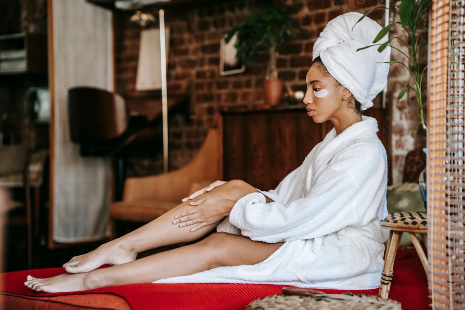 Side view of young female in white robe and white towel on head relaxing on comfortable bed and applying cream on leg