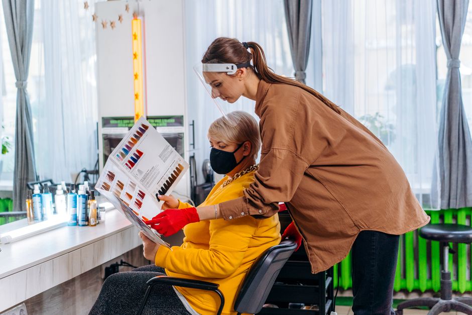 Side view female master in medical mask and gloves showing album with hairstyles images to client on chair in salon