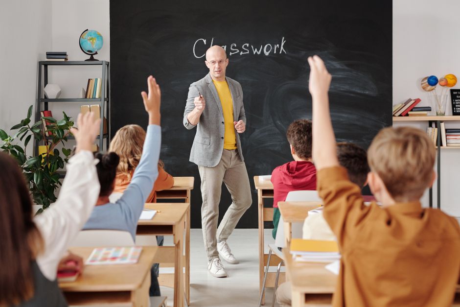 Eine Lehrerin im Austausch mit Schülerinnen und Schülern, die im Klassenzimmer die Hand heben.