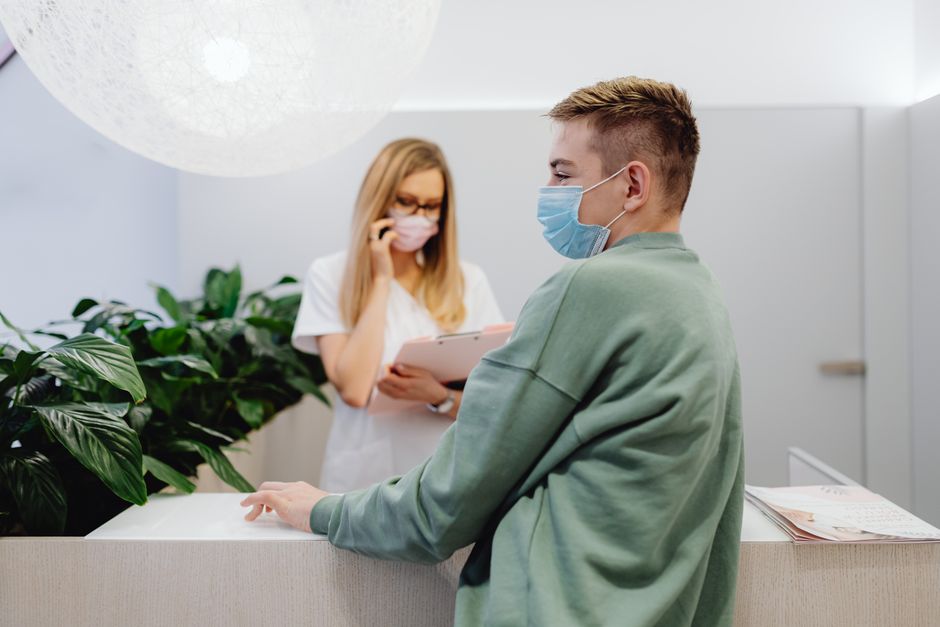 Pacientas ir registratūros darbuotojas klinikoje dėvi veido kaukes, užtikrindami sveikatos saugumą.