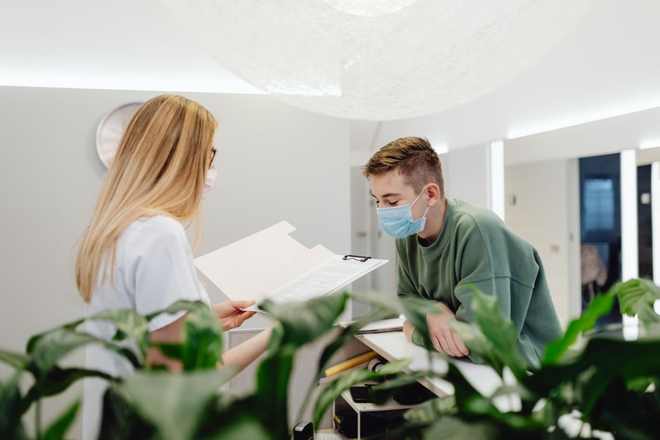 Moteris ir vyras su apsauginėmis kaukėmis kalbasi prie klinikos registratūros stalo patalpoje.